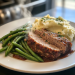 A beautifully glazed homemade meatloaf sliced and served with mashed potatoes and green beans on a dinner plate.